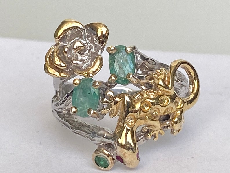 May include: A silver ring with a gold lizard, a gold flower, and green gemstones.