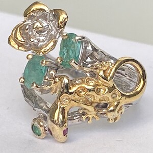 May include: A silver ring with a gold lizard design, a flower, and green gemstones.
