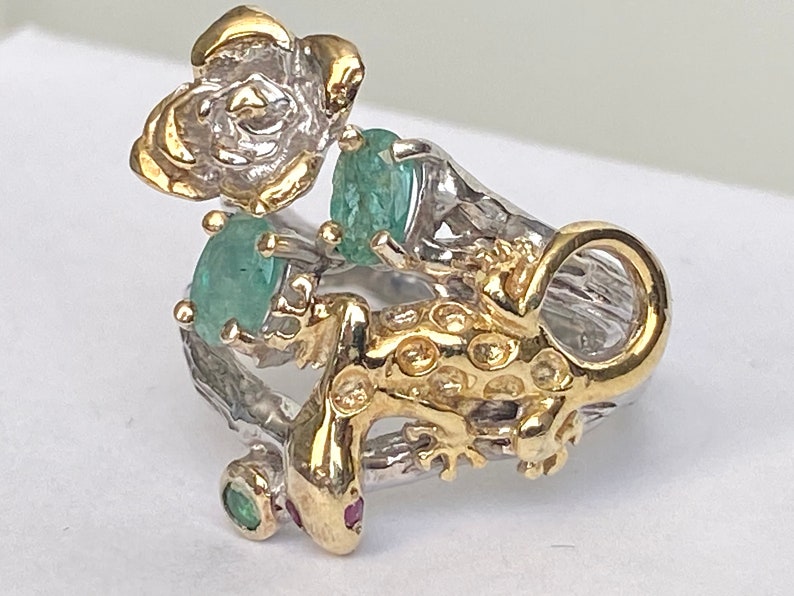 May include: A silver ring with a gold lizard design, a flower, and green gemstones.