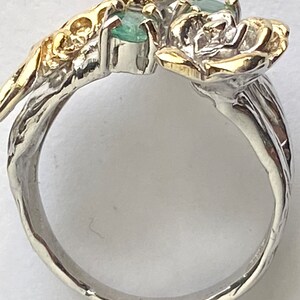 May include: A silver ring with a gold rose design and two emerald green gemstones. The ring is set with a white pearl and has a textured band.