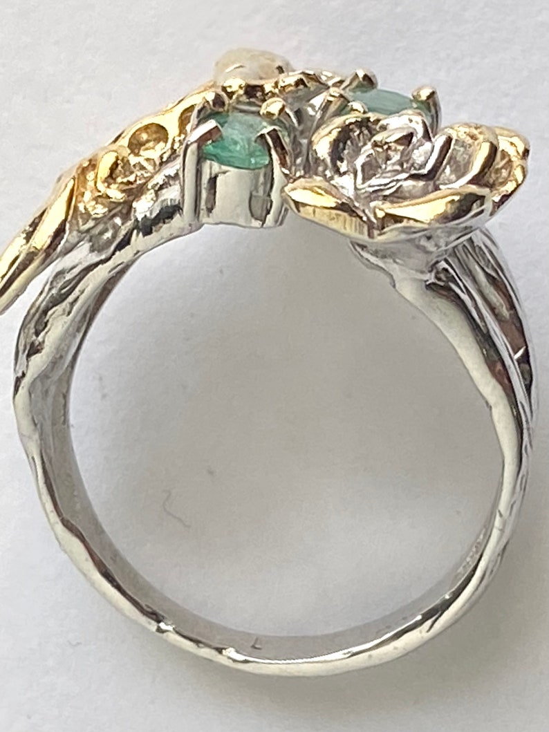 May include: A silver ring with a gold rose design and two emerald green gemstones. The ring is set with a white pearl and has a textured band.