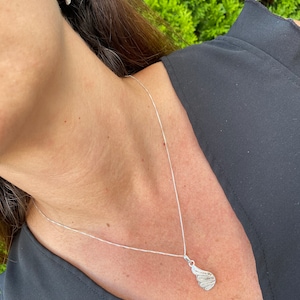 Puede incluir: Un collar de plata con un delicado colgante de ala de filigrana. El collar lo lleva una persona con el pelo oscuro y una camisa negra.