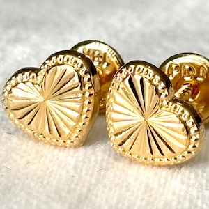 May include: Close-up of two gold heart-shaped stud earrings. The earrings have a textured surface with a sunburst design and a beaded border. The earrings are on a white surface.