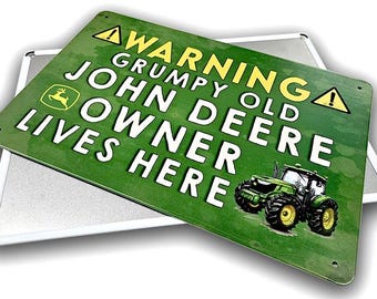 Puede incluir: Un letrero metálico verde con el texto "WARNING GRUMPY OLD JOHN DEERE OWNER LIVES HERE" y un gráfico de tractor John Deere. El letrero tiene un aspecto envejecido y está montado sobre un respaldo de metal plateado.