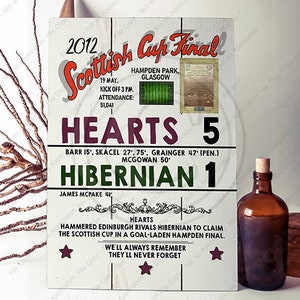 May include: A white wooden sign with the text "2012 Scottish Cup Final" and the results of the game: Hearts 5, Hibernian 1. The sign also includes the date, time, and attendance of the game. The sign is decorated with stars and the text "We'll always remember, they'll never forget."