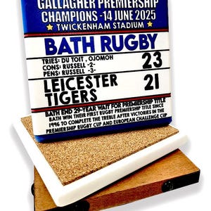 May include: A rectangular coaster with a blue top displaying "GALLAGHER PREMIERSHIP CHAMPIONS - 14 JUNE 2025" and "BATH RUGBY" with the score 23-21 against Leicester Tigers. The coaster rests on a cork and wood base.
