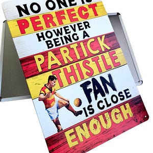 Può includere: Un cartello metallico rettangolare con uno sfondo effetto legno invecchiato. Il cartello presenta il testo "NO ONE IS PERFECT HOWEVER BEING A PARTICK THISTLE FAN IS CLOSE ENOUGH" in lettere colorate e in grassetto. Un calciatore sta calciando un pallone.