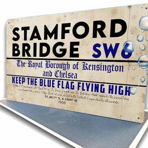 May include: A vintage-style metal sign with the text "STAMFORD BRIDGE SW6" in black and blue. It also reads "The Royal Borough of Kensington and Chelsea" and "KEEP THE BLUE FLAG FLYING HIGH". The sign includes coordinates and the year 1905.
