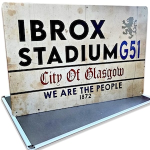 May include: A vintage-style metal sign with the text "IBROX STADIUM G51" in black and blue. The sign also includes "City Of Glasgow" in red and "WE ARE THE PEOPLE 1872" in blue. The sign is mounted on a metal base.
