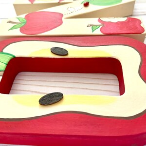 May include: A rectangular wooden frame painted to resemble a sliced apple with a red skin, cream-colored flesh, and two dark brown oval seeds. Behind it, a wooden letter M is decorated with painted red apples and a small ladybug.