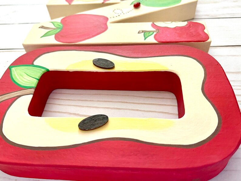 May include: A rectangular wooden frame painted to resemble a sliced apple with a red skin, cream-colored flesh, and two dark brown oval seeds. Behind it, a wooden letter M is decorated with painted red apples and a small ladybug.