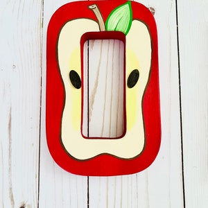May include: A rectangular wooden light switch cover plate painted to resemble a sliced apple. The design features a bright red border, a cream-colored center, two dark brown seeds, and a small green leaf attached to a brown stem at the top.