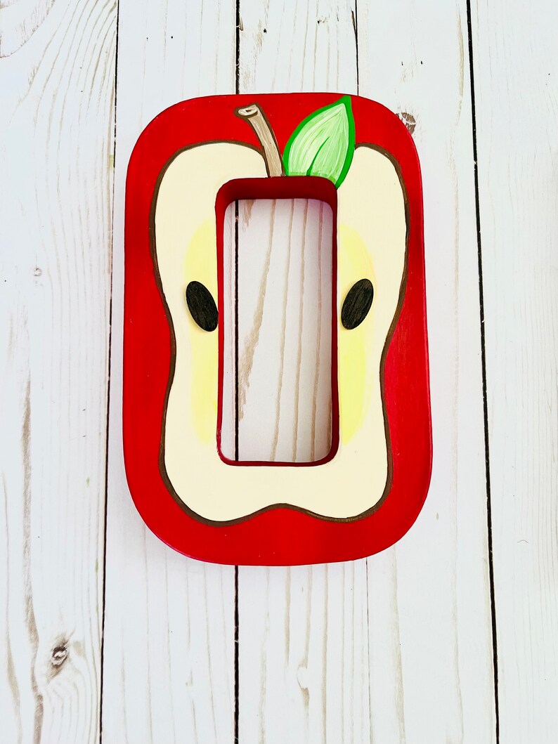May include: A rectangular wooden light switch cover plate painted to resemble a sliced apple. The design features a bright red border, a cream-colored center, two dark brown seeds, and a small green leaf attached to a brown stem at the top.