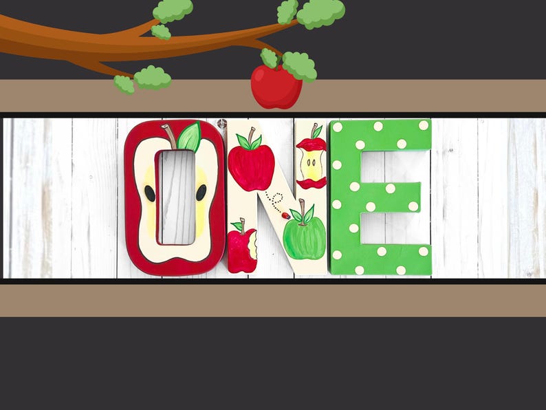 May include: Three decorative wooden letters spelling the word ONE. The O is shaped like a sliced apple, the N features painted red and green apples with a ladybug, and the E is green with white polka dots. The letters are arranged horizontally.