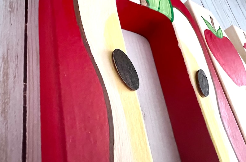 May include: A wooden wall-mounted coat rack featuring a series of painted red apple motifs on a cream-colored background. The rack includes dark brown circular hooks attached to the front, with each apple design accented by green leaves and brown stems.