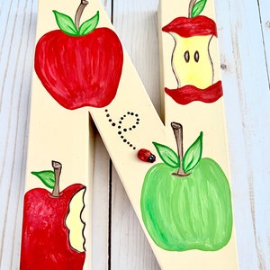 May include: A cream-colored wooden letter N decorated with hand-painted illustrations of two whole apples, one red and one green, and two apple cores. A small three-dimensional ladybug is attached to the surface with a painted dotted trail.