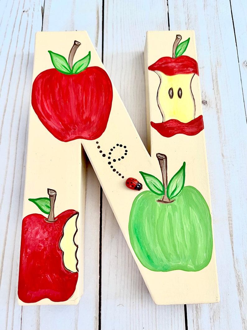 May include: A cream-colored wooden letter N decorated with hand-painted illustrations of two whole apples, one red and one green, and two apple cores. A small three-dimensional ladybug is attached to the surface with a painted dotted trail.