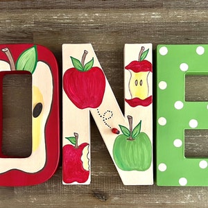 May include: Three-dimensional wooden letters spelling the word ONE. The letter O is red with a sliced apple design. The letter N is cream-colored with painted red and green apples, an apple core, and a ladybug. The letter E is green with white polka dots.