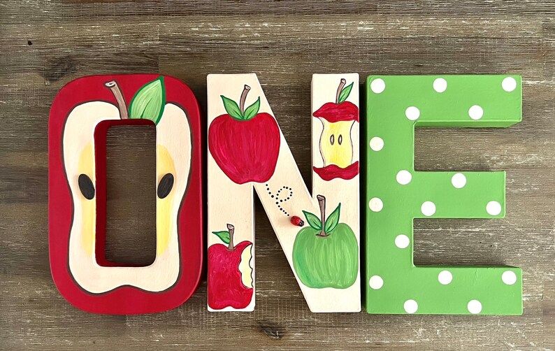 May include: Three-dimensional wooden letters spelling the word ONE. The letter O is red with a sliced apple design. The letter N is cream-colored with painted red and green apples, an apple core, and a ladybug. The letter E is green with white polka dots.