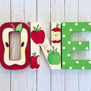 May include: Three large wooden letters spelling the word ONE. The letter O is red and shaped like a sliced apple. The letter N is cream-colored with painted red and green apples and a ladybug. The letter E is green with white polka dots.