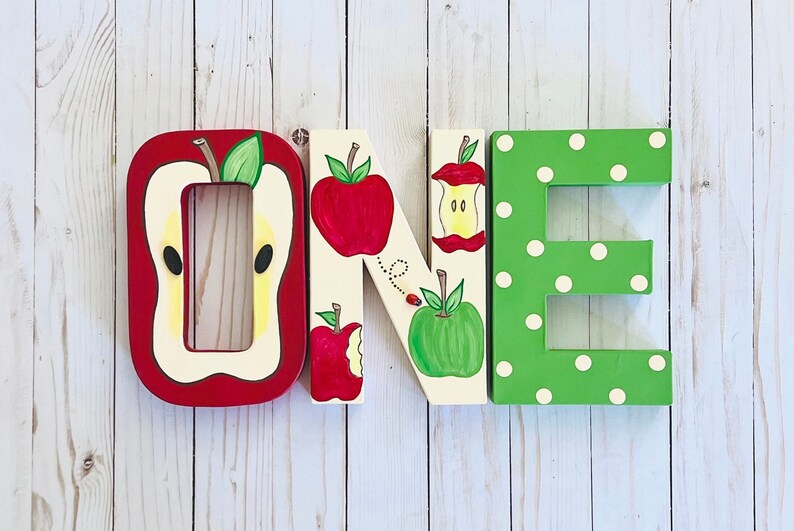 May include: Three large wooden letters spelling the word ONE. The letter O is red and shaped like a sliced apple. The letter N is cream-colored with painted red and green apples and a ladybug. The letter E is green with white polka dots.