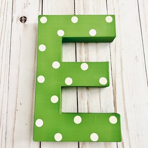 May include: A three-dimensional letter E painted in a vibrant grass green color, featuring a repeating pattern of small, circular white polka dots across its entire front surface. The letter has a blocky, thick structure with clean, straight edges.