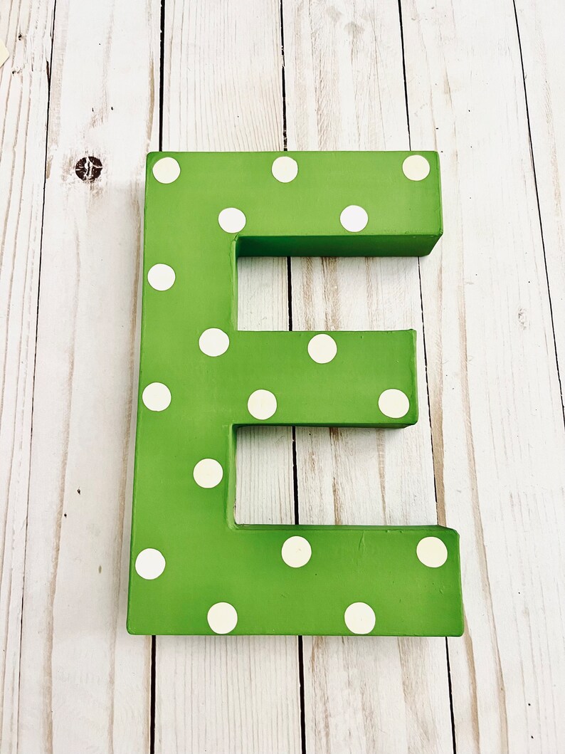 May include: A three-dimensional letter E painted in a vibrant grass green color, featuring a repeating pattern of small, circular white polka dots across its entire front surface. The letter has a blocky, thick structure with clean, straight edges.