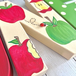 May include: A cream-colored wooden letter N decorated with hand-painted red and green apples, an apple core, and a small three-dimensional ladybug embellishment. A dotted line trail follows the ladybug across the surface of the letter.