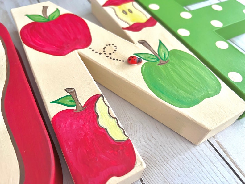 May include: A cream-colored wooden letter N decorated with hand-painted red and green apples, an apple core, and a small three-dimensional ladybug embellishment. A dotted line trail follows the ladybug across the surface of the letter.