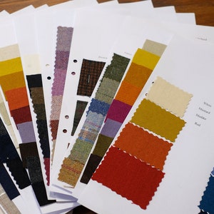 Op de afbeelding: Een stoffenstaalboek met een verscheidenheid aan kleuren en texturen. Het boek bevat stoffenstalen in wit, mosterd, rood, olijf, lichtblauw, petrol en donkerblauw.