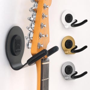May include: A wall-mounted guitar hanger with a black circular base and a black U-shaped support arm holding a light wood guitar neck. Additional hangers are shown in white, gold, and silver finishes, each featuring a black center logo.
