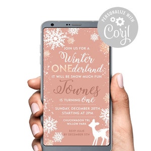Könnte beinhalten: Ein Smartphone zeigt eine rosa Einladung für eine Winter-Geburtstagsfeier. Die Einladung enthält Schneeflocken und ein Reh, sowie die Veranstaltungsdetails und RSVP-Informationen. Das Telefon wird in einer Hand gehalten.