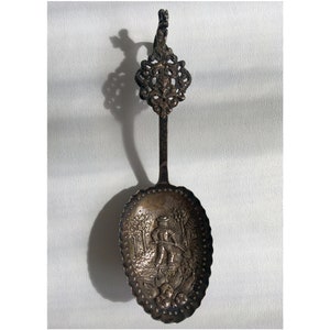 May include: An antique silver spoon with an ornate handle and a detailed scene of a person in a landscape on the bowl.