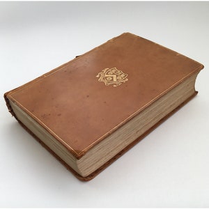 May include: A vintage hardcover book with a brown leather cover. The cover features a gold embossed emblem and gold-gilded edges. The book is closed and appears to be well-preserved, suggesting it is an antique or collectible item.