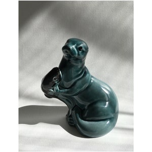 May include: A teal blue ceramic figurine of an otter holding a fish in its paws. The otter is sitting on its haunches with its tail curled around its body.
