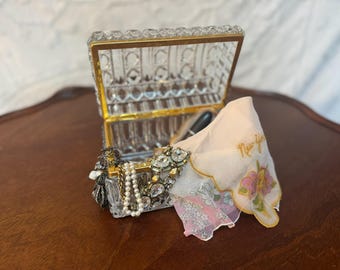May include: A clear, rectangular jewelry box with a gold-colored trim, open to reveal jewelry and a handkerchief. The handkerchief is white with pink floral accents and the text "New York".