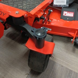 May include: Close-up of an orange and black lawn mower. The mower has a black wheel and a black cylindrical component attached to the orange frame. The tire is black and has a tread pattern. The mower is on a wooden floor.