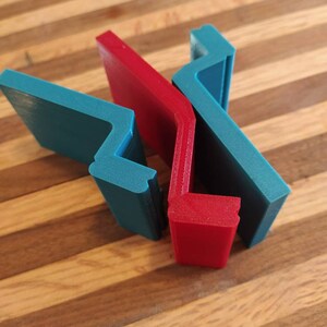 May include: Two 3D printed phone stands, one red and one blue, are stacked on top of each other. The stands are shaped like a right angle with a curved edge.