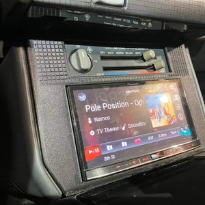 May include: A black car dashboard with a Pioneer car stereo system playing the "Pole Position - Opt" soundtrack. The screen displays the song title, artist, and album art. The stereo has a black and silver finish and features buttons for volume, track selection, and other functions.