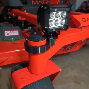 Può includere: Un tosaerba rosso con una luce a LED nera montata su una staffa in metallo nero. La luce ha sei lampadine LED individuali. Il tosaerba ha un parafango in plastica nera e un pneumatico nero.