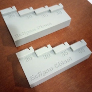 Op de afbeelding: Twee grijze kunststof linialen met markeringen voor verschillende maten. De bovenste liniaal is gemarkeerd met "Eclipse Plane" en heeft markeringen voor 25, 30, 35 en 38. De onderste liniaal is gemarkeerd met "Eclipse Chisel" en heeft markeringen voor 25, 30, 35 en 40.