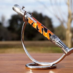 May include: A silver pen with a black and orange flame design on the barrel. The pen is displayed on a silver stand.