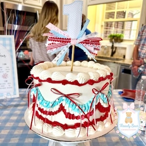 K&ouml;nnte beinhalten: Eine runde Torte mit wei&szlig;er Glasur, verziert mit roten und hellblauen aufgespritzten Icing-Girlanden und d&uuml;nnen roten B&auml;ndern. Ein Tortenstecker in Form einer Eins aus Stoff kr&ouml;nt die Torte, geschm&uuml;ckt mit roten, wei&szlig;en und blauen B&auml;ndern.