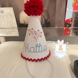 K&ouml;nnte beinhalten: Ein konischer Partyhut aus wei&szlig;em Stoff mit dezentem roten Punktmuster, gekr&ouml;nt von einem gro&szlig;en roten Wollpompon. Der Hut ist mit zwei Wunderkerzen und dem Namen Hattie in blauer Schreibschrift bestickt und hat einen roten Zackenlitzenrand.