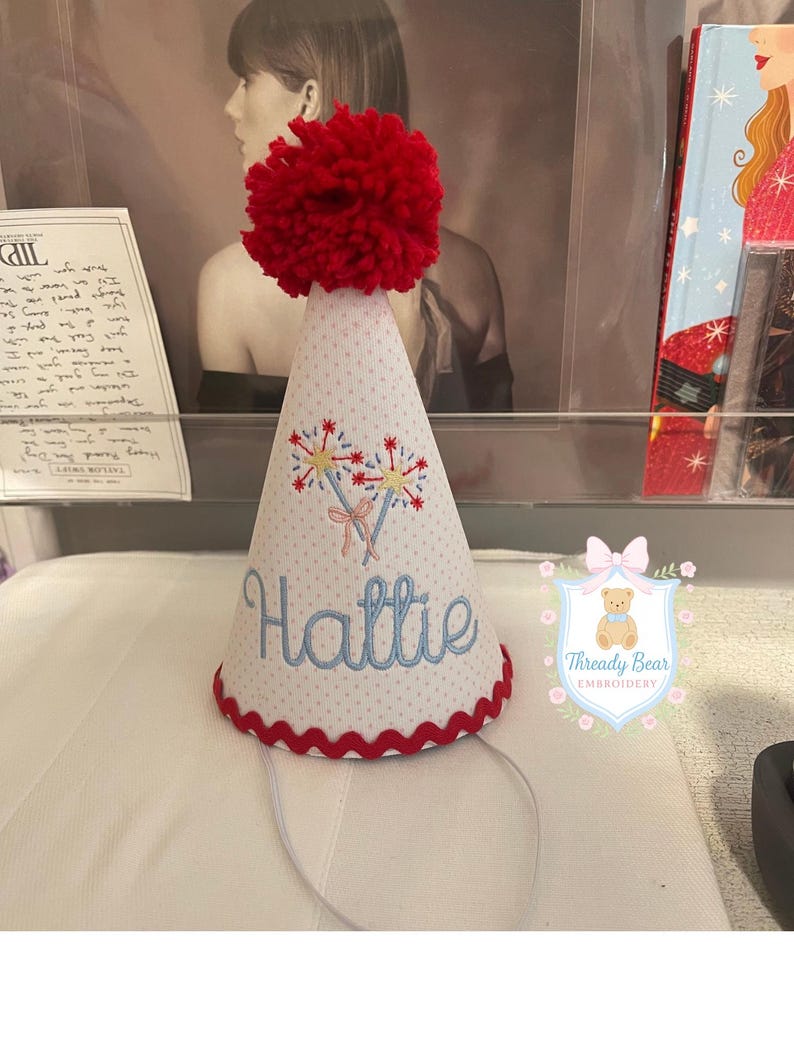 K&ouml;nnte beinhalten: Ein konischer Partyhut aus wei&szlig;em Stoff mit dezentem roten Punktmuster, gekr&ouml;nt von einem gro&szlig;en roten Wollpompon. Der Hut ist mit zwei Wunderkerzen und dem Namen Hattie in blauer Schreibschrift bestickt und hat einen roten Zackenlitzenrand.