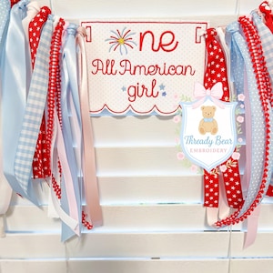 K&ouml;nnte beinhalten: Ein Stoffbanner mit einem zentralen rechteckigen Paneel mit gewelltem Rand, bestickt mit dem roten Text 'one All American girl' und einem Feuerwerksmotiv. Das Banner ist mit verschiedenen roten, wei&szlig;en und blauen B&auml;ndern verziert.