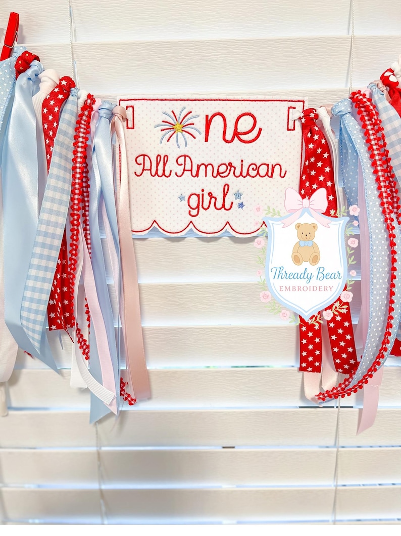 K&ouml;nnte beinhalten: Ein Stoffbanner mit einem zentralen rechteckigen Paneel mit gewelltem Rand, bestickt mit dem roten Text 'one All American girl' und einem Feuerwerksmotiv. Das Banner ist mit verschiedenen roten, wei&szlig;en und blauen B&auml;ndern verziert.