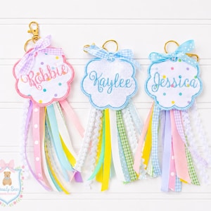 May include: Three personalized keychains with cloud-shaped embroidered name tags. Each tag has a different name in cursive script: Robbie, Kaylee, and Jessica. The keychains feature colorful ribbons and a gold-tone clasp.