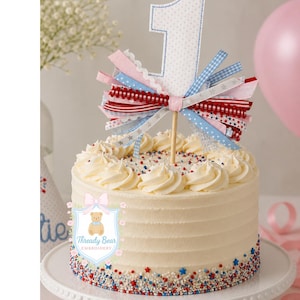 K&ouml;nnte beinhalten: Ein wei&szlig; glasierter Kuchen mit einem Stoff-Tortenstecker in Form der Zahl Eins mit blauem Punktmuster. Der Tortenstecker ist mit einem B&uuml;ndel aus rot-wei&szlig;-blau gemusterten B&auml;ndern verziert. Der Kuchenboden ist mit rot-wei&szlig;-blauen Streuseln bedeckt.