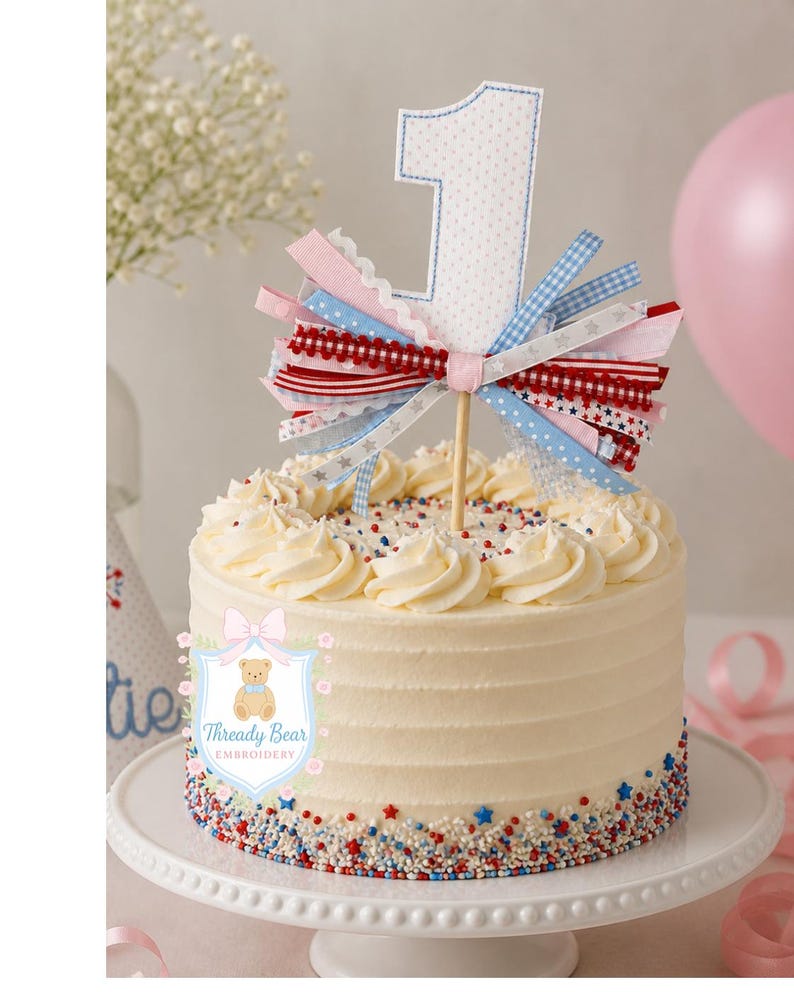K&ouml;nnte beinhalten: Ein wei&szlig; glasierter Kuchen mit einem Stoff-Tortenstecker in Form der Zahl Eins mit blauem Punktmuster. Der Tortenstecker ist mit einem B&uuml;ndel aus rot-wei&szlig;-blau gemusterten B&auml;ndern verziert. Der Kuchenboden ist mit rot-wei&szlig;-blauen Streuseln bedeckt.